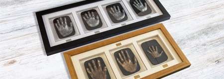 Framed handprint plaques display rows of metallic hand impressions; one black frame holds four, one wooden frame holds three, lying on a whitewashed wooden tabletop.