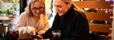 Two people smile while looking at a smartphone; one points at the screen. They sit at a cafe table with two dark sodas, warm lighting, and wooden booth seating.