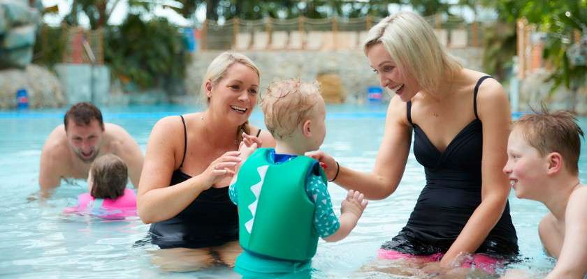 Child wearing a green flotation vest stands and splashes; two adults guide and smile while other children and an adult swim nearby in a shallow indoor pool surrounded by tropical decor.