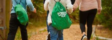 Hikers carry green drawstring backpacks, walking away along a gravel forest path. Surrounding trees and foliage line the trail. Visible bag text: "FOREST DISCOVERY KITS".