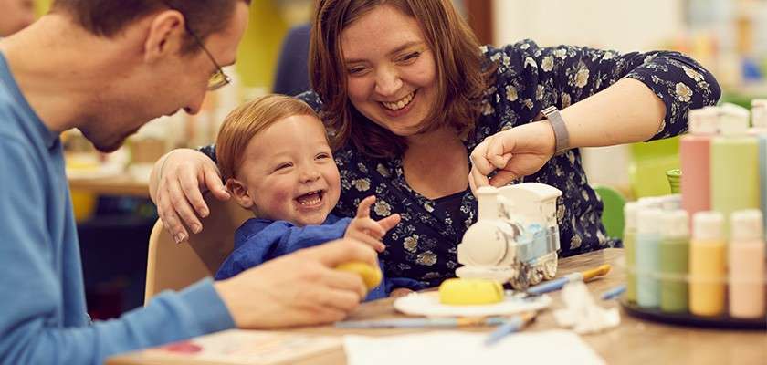 Child laughs while painting a ceramic train with two adults at a table, surrounded by paint bottles, brushes, sponges, and papers in a bright indoor craft studio.