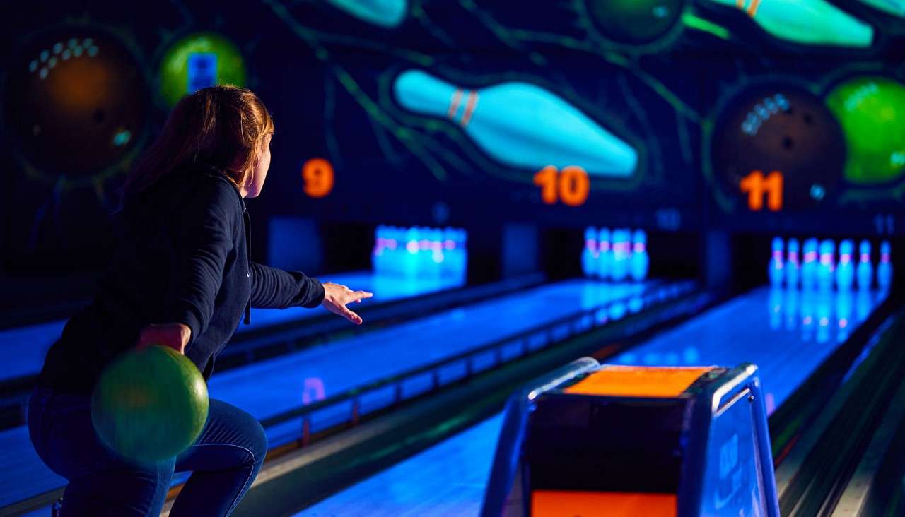 Bowler with green ball releases it down a neon-lit lane, arm extended. Glowing pins wait ahead; surrounding lanes and ball return shine under blacklight. Visible text: 9, 10, 11.