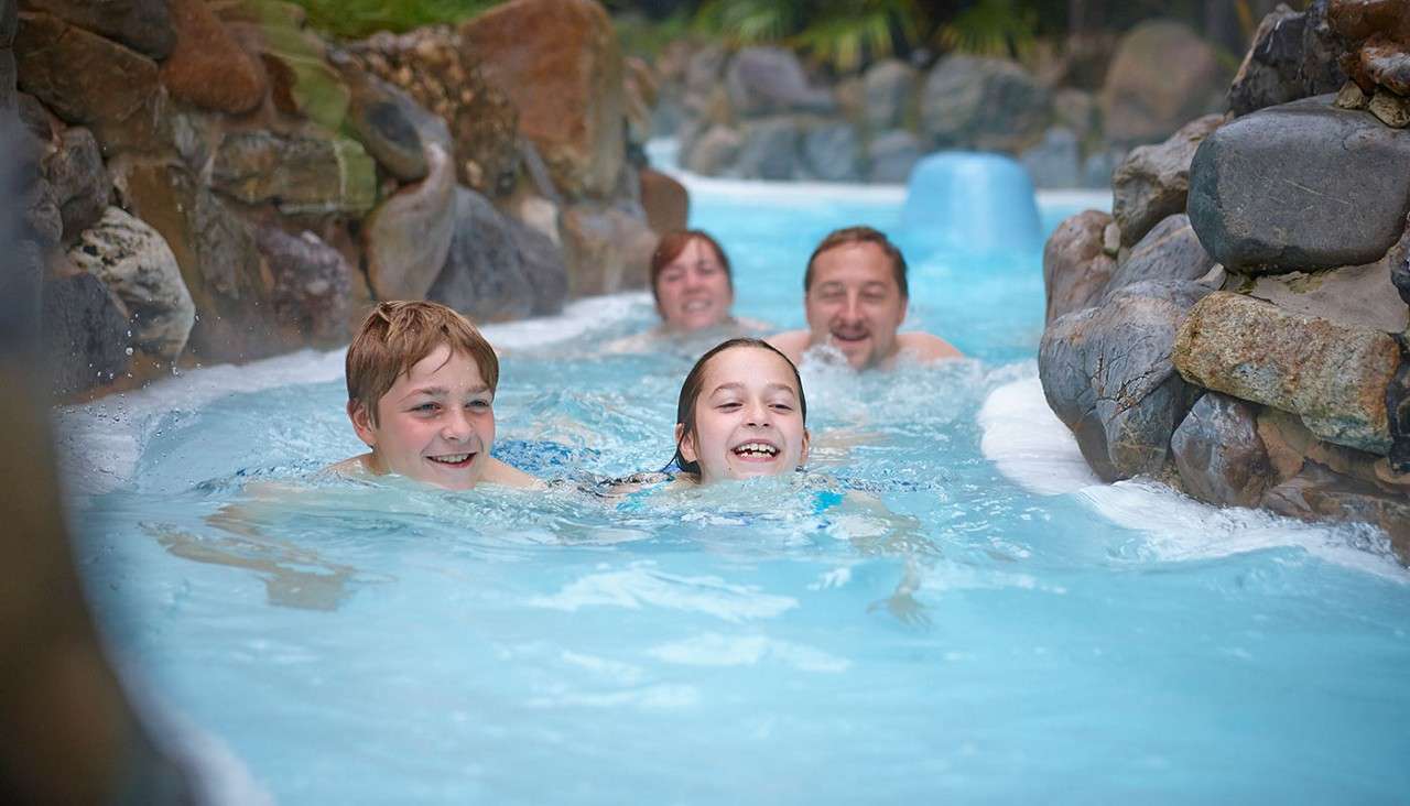 Two children swim ahead while two adults follow, smiling, in a shallow, blue, rock-lined lazy river at a water park, water bubbling along a narrow channel.