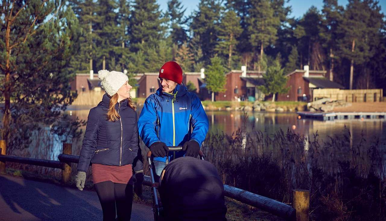 Couple with a stroller walk and chat on a paved lakeside path, bundled in winter jackets and hats. Behind them, wooden cabins face a calm pond amid tall evergreen trees. Text: Berghaus.