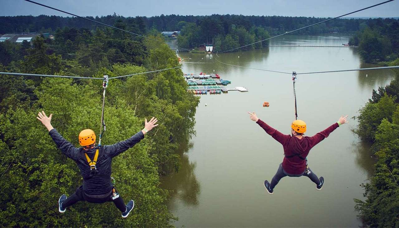 Two helmeted zipliners glide with arms outstretched on parallel cables, suspended high above a wide lake. Dense green forest lines the water, with docks and small boats ahead under an overcast sky.