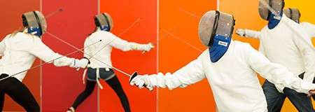Fencers lunge and parry with foils, arms extended mid-thrust. They wear masks and white jackets. Context: a practice session indoors against a vibrant orange-to-red gradient wall.