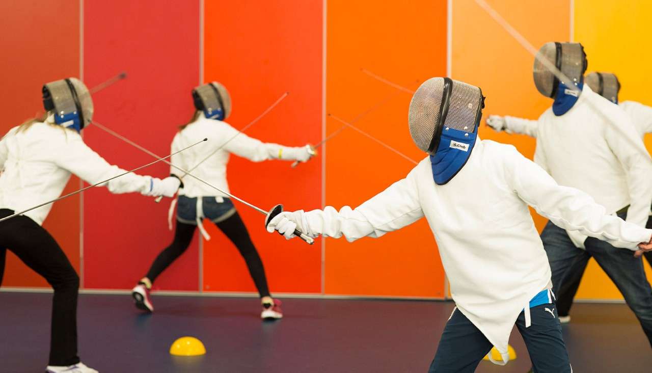 Fencers lunge with foils, practicing footwork; several people in masks and white jackets train in a gym against a gradient orange wall. Visible text: PUMA; Club.