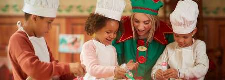 Children in chef hats decorate cookies with icing alongside an adult dressed as a holiday elf, in a warm, festive kitchen with wooden walls and garland.
