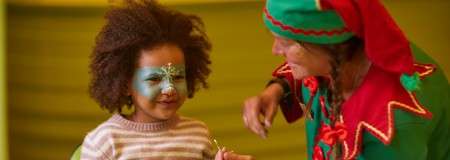 Child with blue-and-white face paint smiles while being painted by a person in a red-and-green elf costume; they sit indoors against a green background, suggesting a festive event.