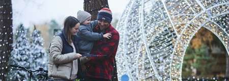 Two adults hold a child and view a tablet, standing outdoors beside a large glowing light arch; snow-dusted trees, winter clothing, and a parked bicycle create a festive park setting.
