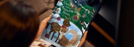 Children’s book—being held—displays a cartoon fawn and fox in a snowy, decorated forest on a dark couch. Visible text: “Clover,” “Dancing Sugar,” and smaller: “By …” (rest unreadable).