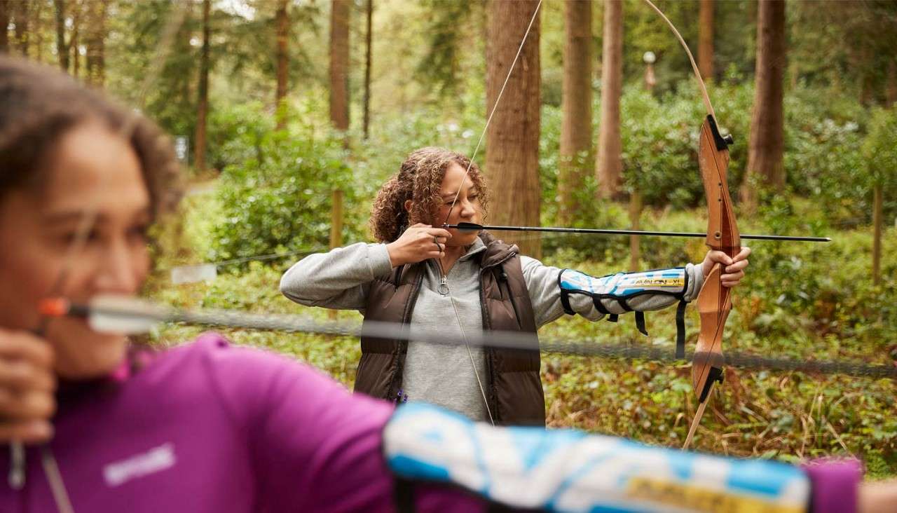 Archer draws a recurve bow, anchoring the string at her cheek. Another archer is blurred in the foreground. Context: outdoor archery range set among tall trees and greenery.