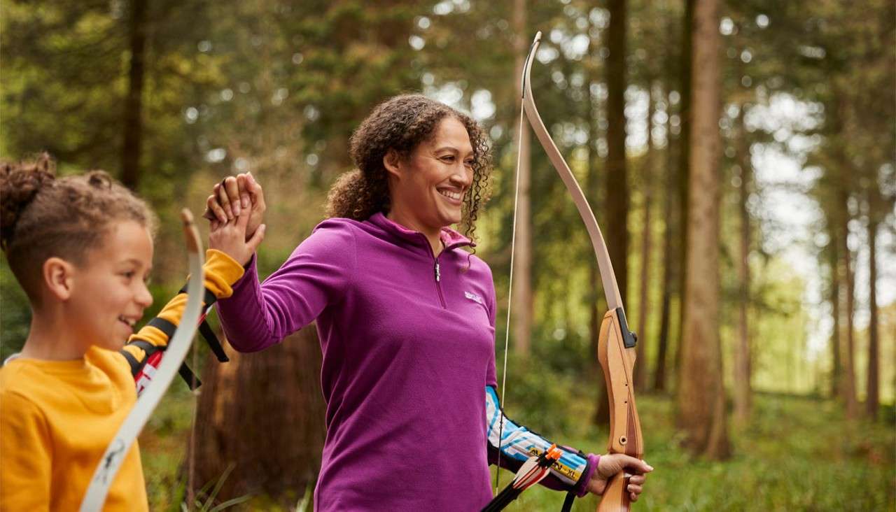Smiling woman archer high-fives a child, holding a wooden recurve bow. Both wear armguards and carry bows while standing in a sunlit forest clearing with tall trees.