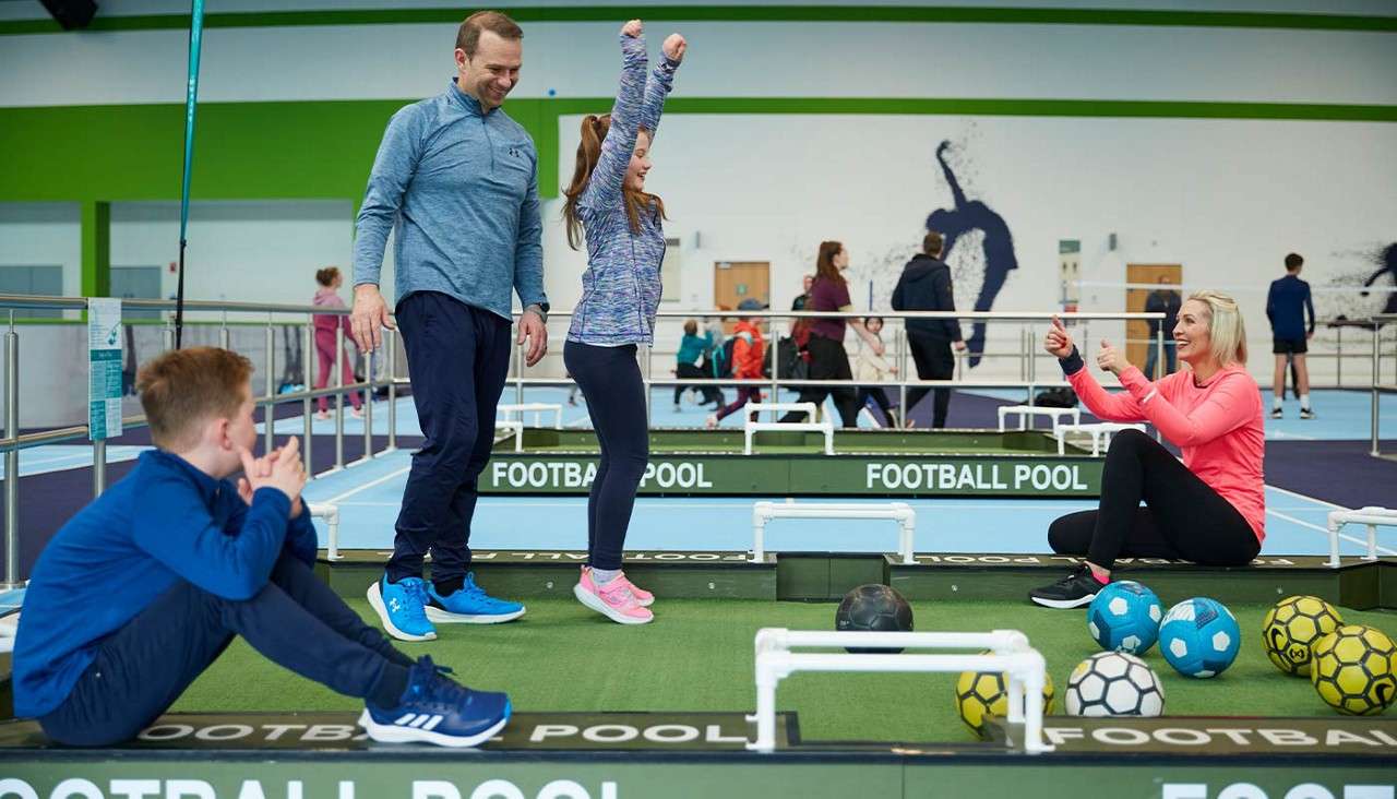 Girl raises arms celebrating; parents and brother watch as they play football pool on mini green pitches with colored balls in an indoor sports hall. Text: FOOTBALL POOL, FOOTBALL POOL, FOOTBALL POOL.