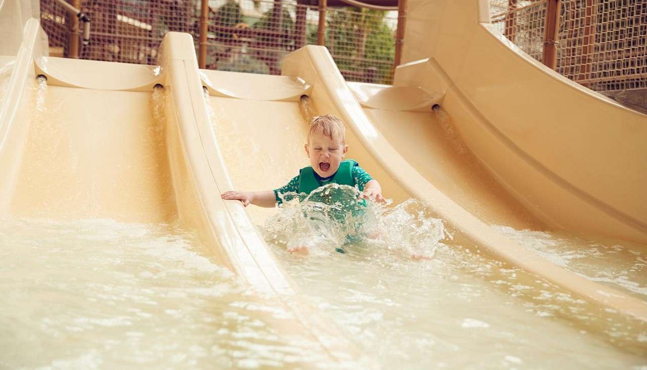 Child slides into shallow pool, splashing water with feet and hands, mouth open, wearing green swim shirt, between beige water slides and netted play structures in an indoor water park.