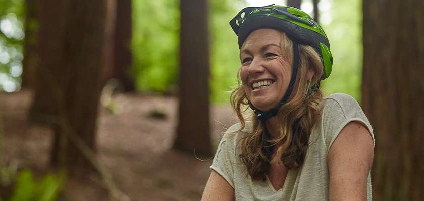 A person in a green bicycle helmet smiles, pausing during a ride; relaxed posture and light-gray shirt amid tall forest trees with soft sunlight filtering through the green canopy.