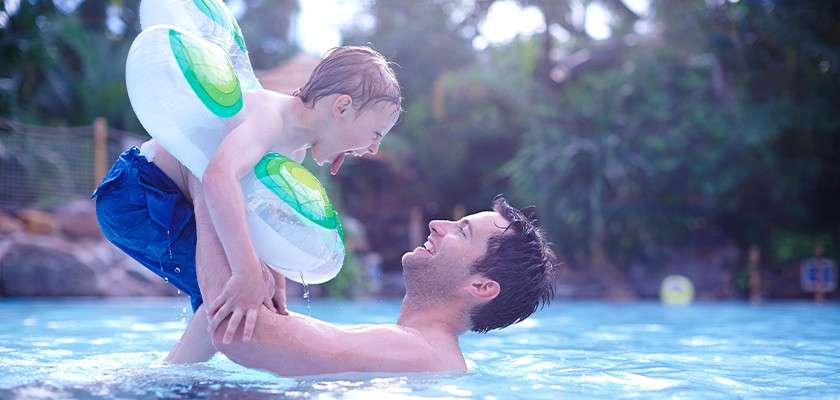 Child with inflatable arm rings laughs while an adult lifts them above the water, splashing playfully in a shallow outdoor pool amid tropical greenery.