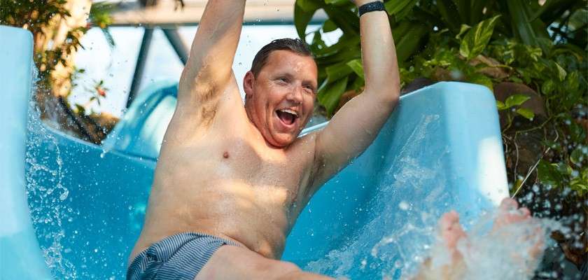 Man slides down a blue water slide, arms raised and smiling, splashing water; tropical plants and an indoor structure around suggest a lively aquatic park setting.