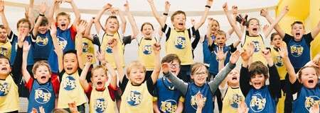 Children cheer with arms raised, smiling widely. They wear matching yellow and blue shirts with cartoon logos, standing closely together in a bright indoor play or sports setting.