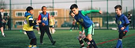 Children playing soccer dribble and chase the ball, wearing blue and yellow training bibs. They run on an artificial-turf field, with cones, a fence, and school buildings in the background.