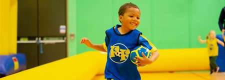 Child runs while holding a small ball, smiling; inside a colorful indoor play area with an inflatable yellow barrier, bright green walls, and another child plus an adult blurred in the background.