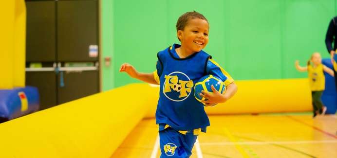 Smiling child runs, clutching a blue ball, in an indoor gym. Yellow inflatable barriers edge the court while colorful walls and another child in back suggest a playful sports session.