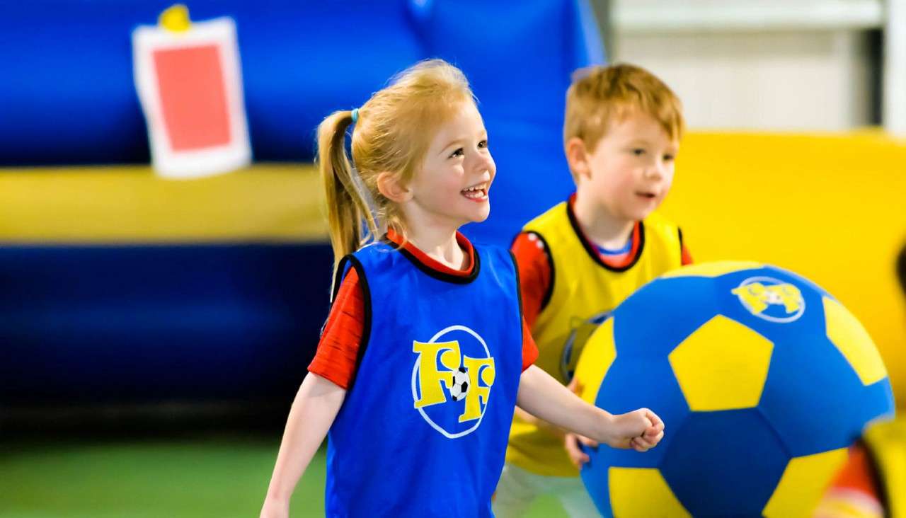 Smiling young girl runs while playing; behind her, a boy rolls a large blue-and-yellow ball in an indoor play area. Her blue vest and the ball display the text “FF.”