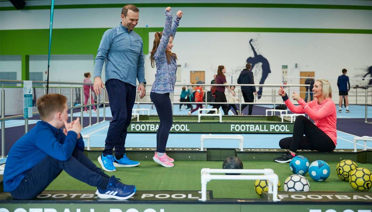 Family plays football pool; girl celebrates with raised arms while mother cheers and father watches; boy sits nearby. Indoor sports hall with lanes and soccer balls. Text: FOOTBALL POOL FOOTBALL POOL FOOTBALL POOL.