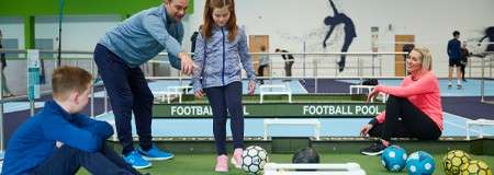 Girl prepares to kick a soccer ball as a man guides her, while a boy and a woman watch; indoor sports center with mini tables and wall silhouettes. Text: FOOTBALL POOL FOOTBALL POOL.