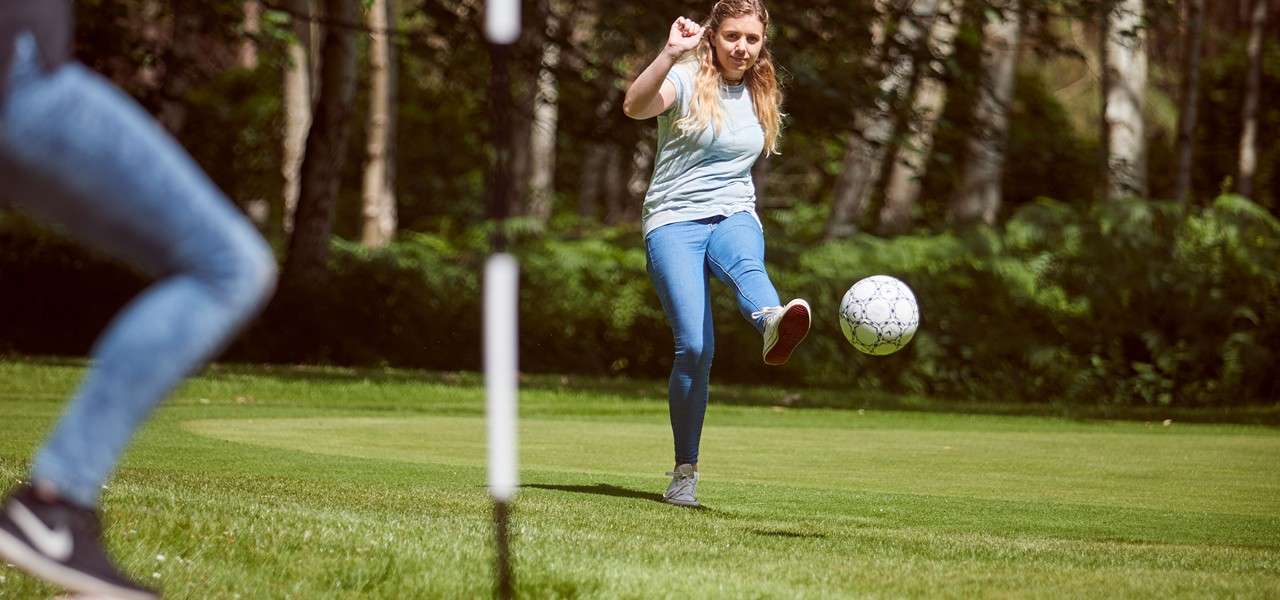 Woman kicks a soccer ball, sending it forward. She stands on a manicured grassy field with a flagpole and trees in the background; another player moves in the foreground.