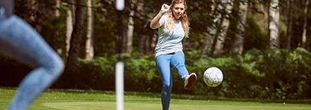 Person kicks a soccer ball toward a flagstick on a grassy course, suggesting footgolf, while another person watches; trees and shrubs surround the open, sunlit park.