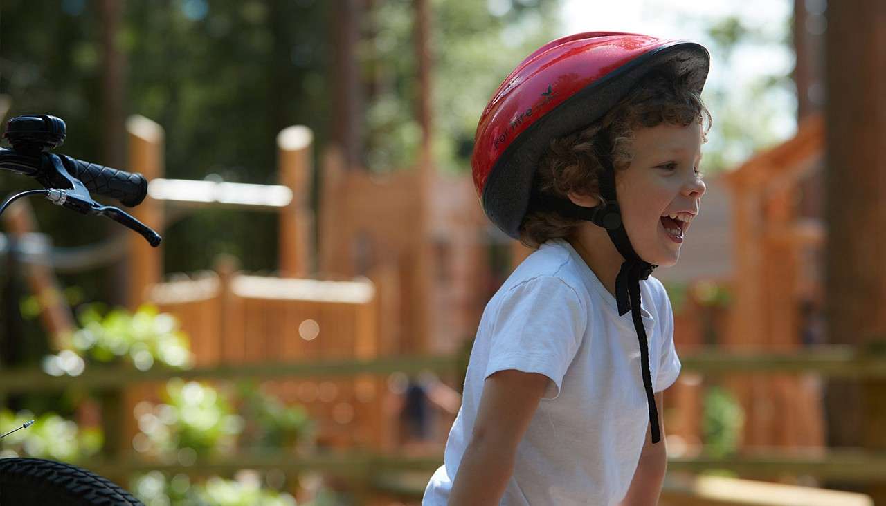 Child wearing a red bike helmet laughs, facing right. Nearby bicycle handlebar in foreground. Sunlit outdoor playground with wooden structures and trees blurred in background.