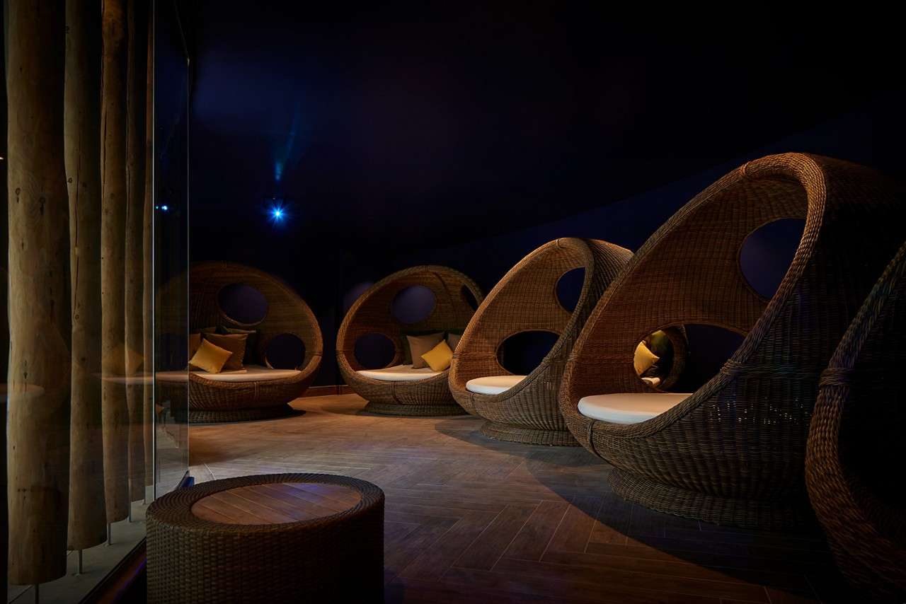 Egg-shaped wicker pod chairs sit empty in a dim spa lounge, with cushions inside circular cutouts, wood-tile flooring, a glass partition of vertical logs, and subtle blue lighting.