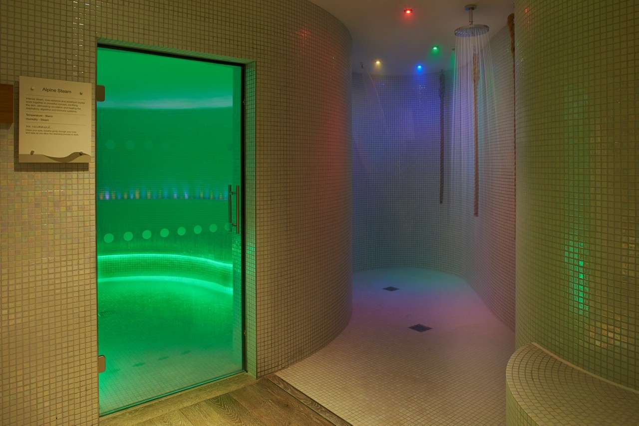 Steam-room door glows green, revealing a curved tiled chamber; adjacent rain shower streams beneath blue, red, and green ceiling lights in a spa hallway. Text: “Alpine Steam”.