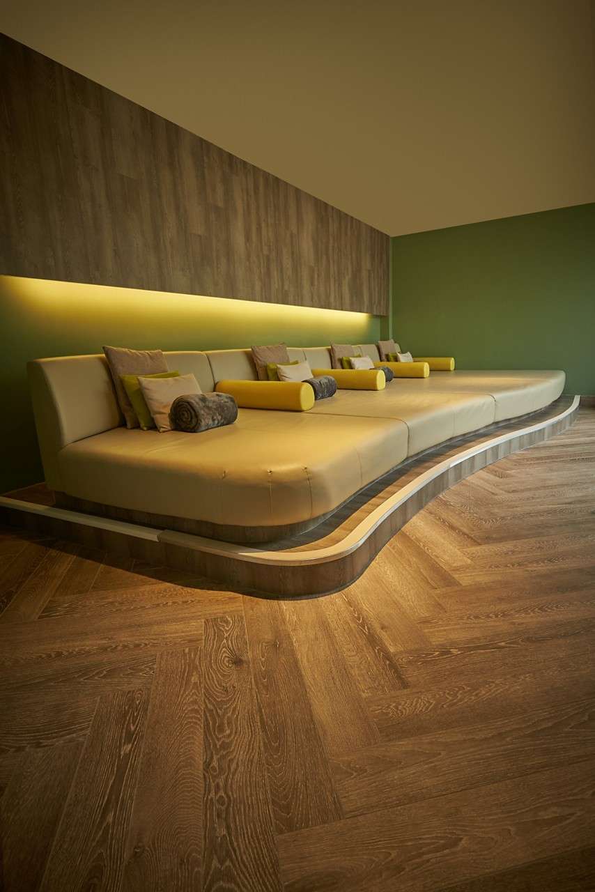 Curved, oversized lounge sofa extends along a wall, topped with pillows and cylindrical bolsters; in context of green walls, wood paneling, underlit strip, and herringbone hardwood floor.