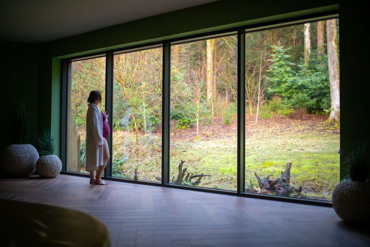 Person stands barefoot, holding their belly, gazing out a wall of floor-to-ceiling windows toward a mossy forest clearing, indoors with wooden floor and large planters.