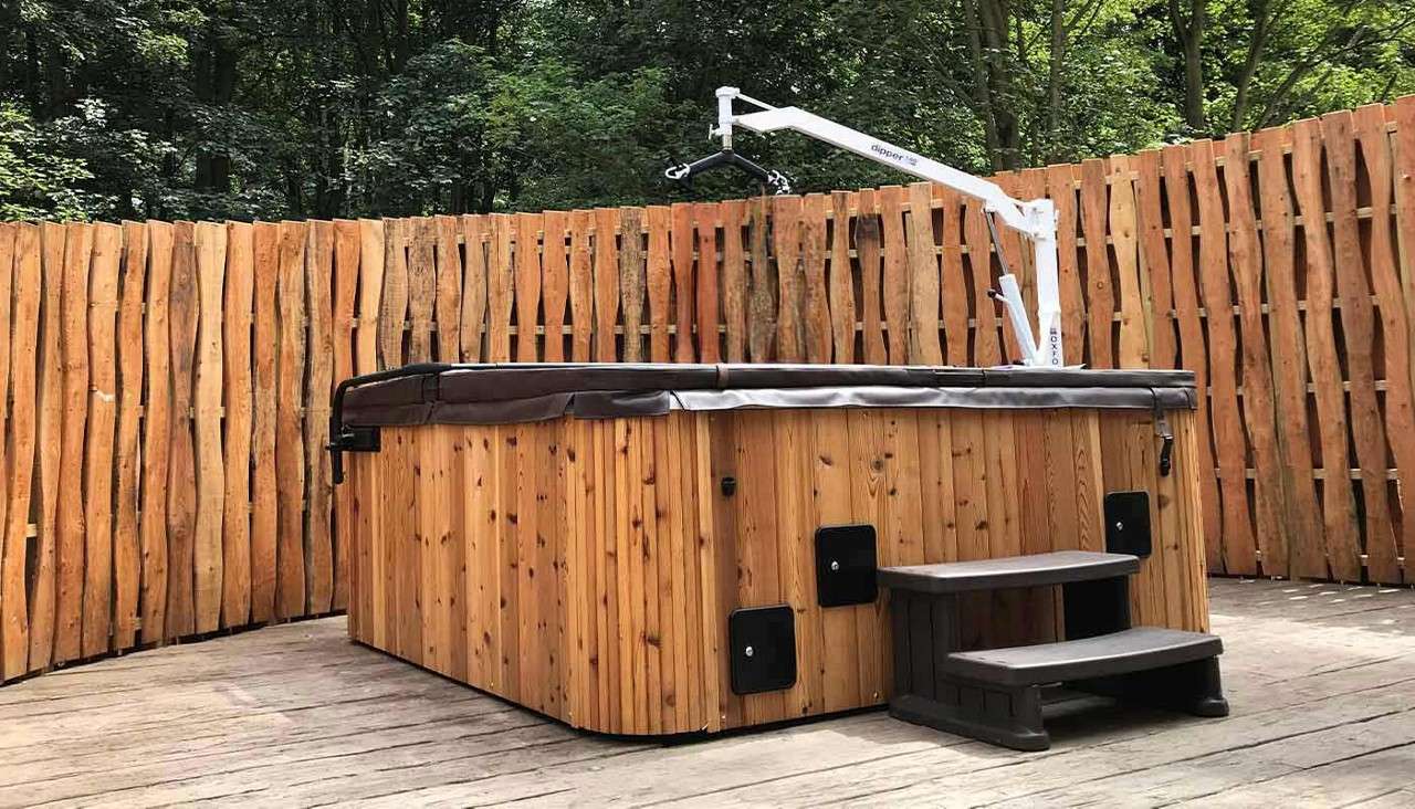 Wooden hot tub with steps stands idle; a white lifting hoist extends above it; surrounded by a tall rustic slatted fence on a deck in a forest. Text: “dipper”, “OXFORD”.