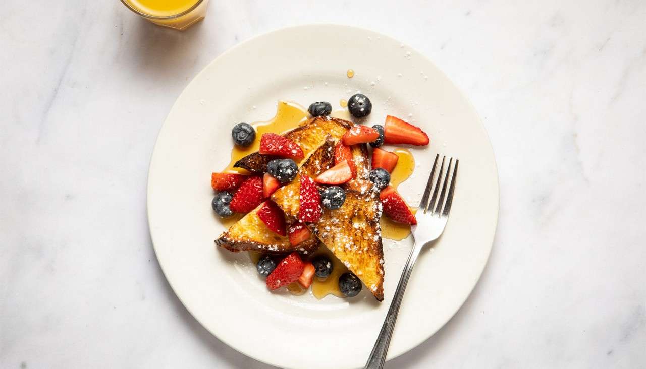 French toast slices topped with strawberries, blueberries, syrup, and powdered sugar; a fork rests on the plate, with a small glass of orange juice nearby on a white countertop.