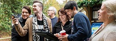 Group of adults laugh while checking a smartphone and tablet; one holds a red coffee cup. In a leafy outdoor setting near an info board, they wear jackets and scarves.