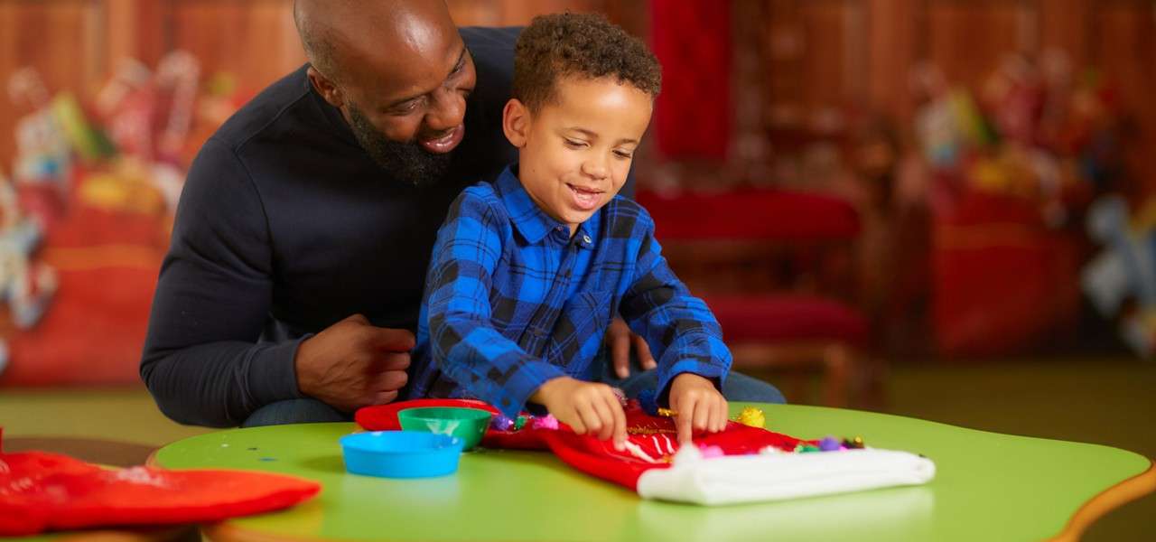 Child decorates a red fabric piece with craft items at a green table, guided by an adult; bowls of supplies nearby; indoor room with blurred red chairs and festive colors.