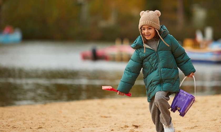 Child runs, holding a red shovel and purple bucket on a sandy beach, bundled in a teal puffer coat and bear-ear knit hat, with water and blurred boats behind.