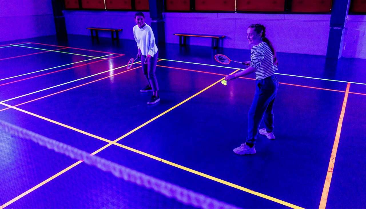 Two people play badminton, rallying a neon shuttlecock, on an indoor court lit with blacklight; glowing yellow-orange court lines, visible net in foreground, benches and walls dimly lit behind.