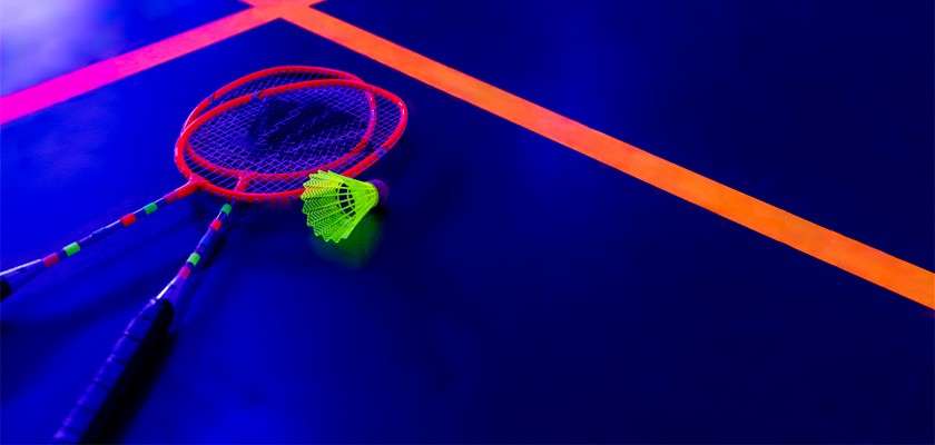 Badminton rackets lie crossed with a neon green shuttlecock resting beside them; context: a dark indoor court glows under blacklight, with bright orange boundary lines on the floor.