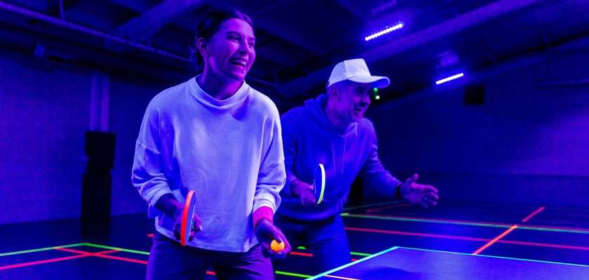 Two people play table tennis, smiling while holding paddles and a ball under blacklight. Neon court lines and violet lighting illuminate an indoor sports hall with overhead fixtures.