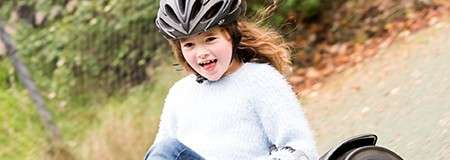 Child in a black helmet rides a bicycle, smiling with mouth open; outdoor setting on a paved path with motion-blurred greenery and fallen leaves in the background.