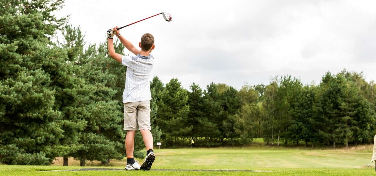 Young golfer completes a driver swing, arms raised. He stands on a tee box facing a fairway, with lush trees bordering the course under a bright, cloudy sky.