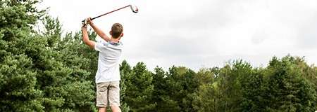 Golfer swings a club in follow-through, viewed from behind; standing on grass, surrounded by dense trees beneath an overcast sky.