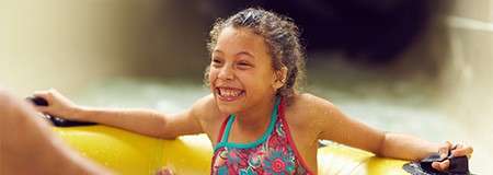 Smiling child rides a yellow inflatable tube, gripping handles, splashing through water in an indoor waterpark or pool, with warm lighting and motion suggesting a gentle slide or current.
