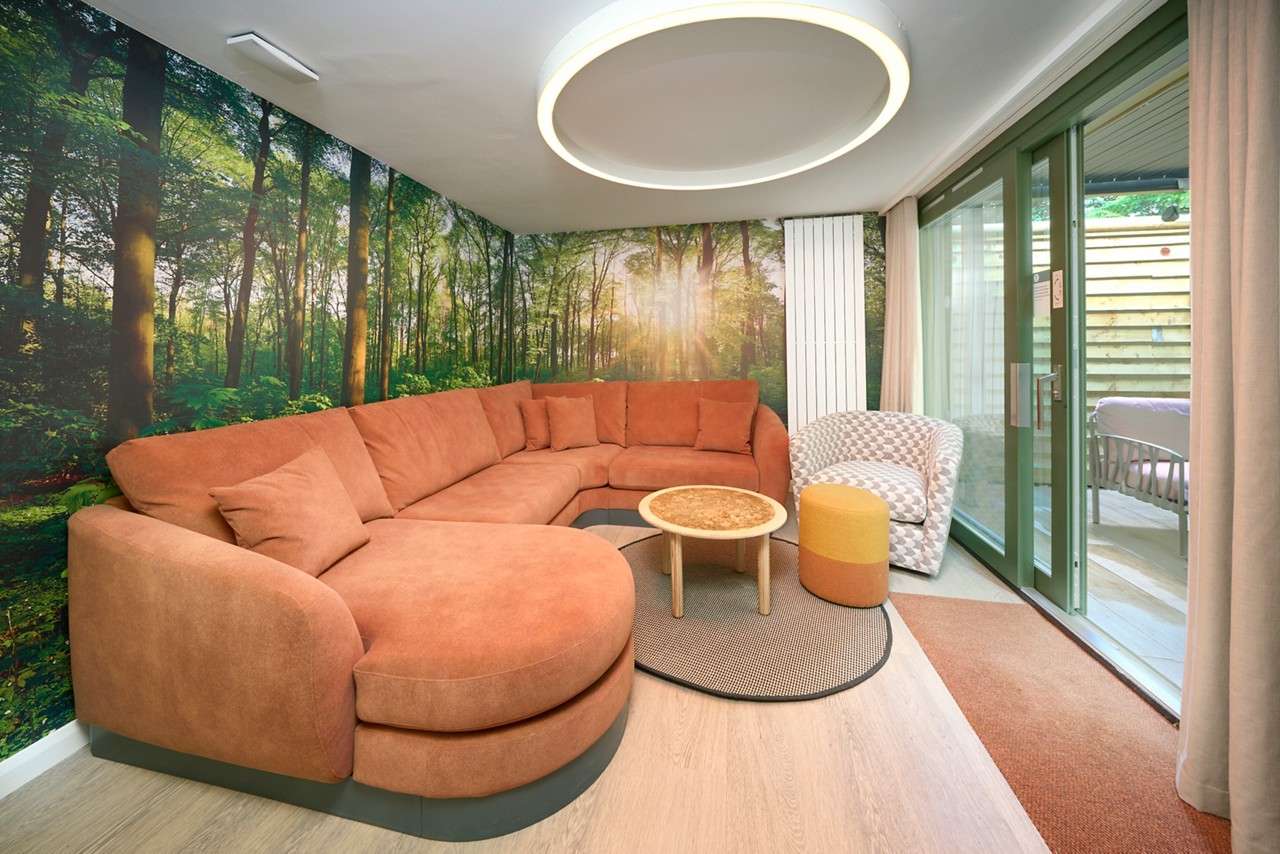 Curved orange sectional sofa surrounds a round coffee table, beneath a circular ceiling light, in a living area with a forest mural and green-framed sliding glass doors to a patio.