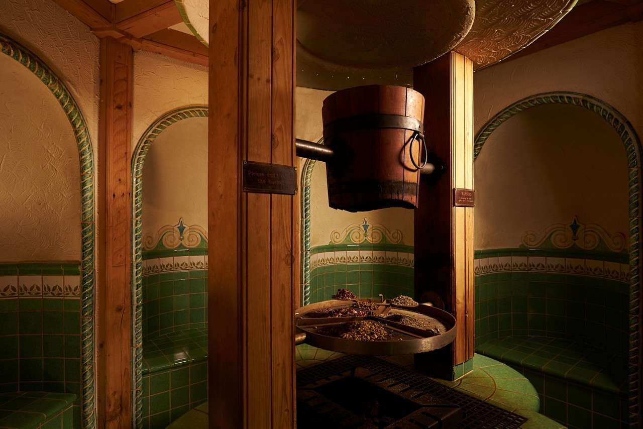 Wooden bucket hangs from beams, dripping over a circular tray of herbs and spices, within an ornate green-tiled steam room with arched alcoves and benches, dimly lit for spa-like ambiance.